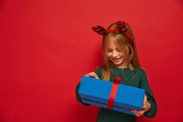 トナカイヘアバンドで面白い子供 女の子 を笑っています クリスマスプレゼントを手に持って クリスマスのコンセプト 赤の背景で撮影 — ストック写真