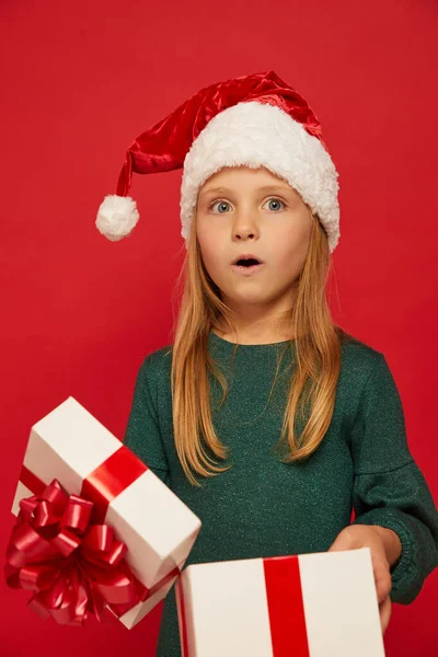 手拿着圣诞礼品盒 戴着圣诞老人红礼帽的笑着顽皮的孩子 圣诞节的概念在红色背景上射击N 图库图片