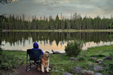 A woman in camping chair sitting on the bank of a calm mountain lake during a gorgeous sunset with her golden retriever dog.  The trees of the  verdant forest are reflected off the water enhancing the serenity of the moment.