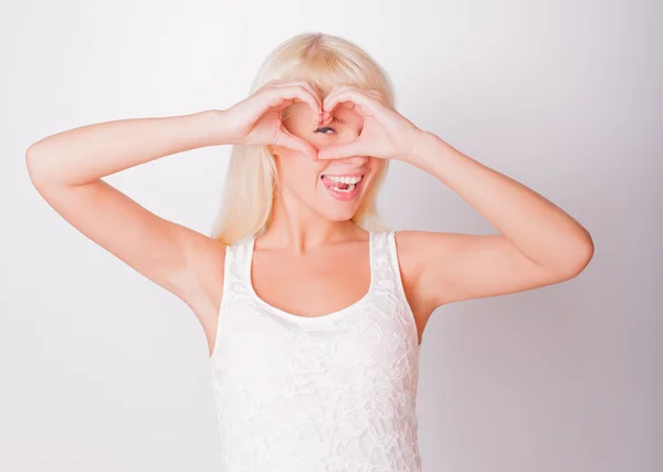 The beautiful fresh blonde looks in a shot through fingers which made heart and smiles — Stock Photo, Image