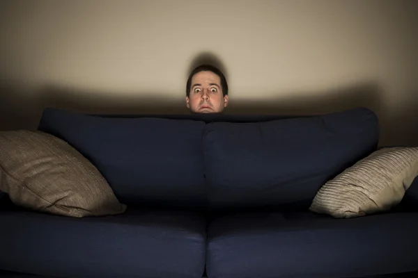 Frightened man peeks over a couch while watching TV — Stock Photo, Image