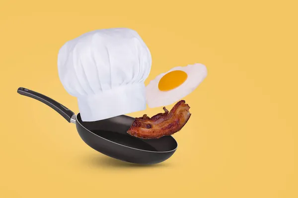最小的想法与油锅 厨师的帽子 飞煎蛋和熏肉的黄色背景 创意食品概念 适用于广告 免版税图库图片