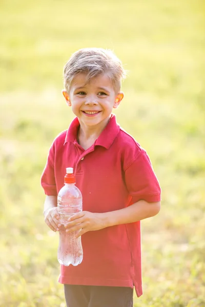 Happy boy with bottle of water on natural background on sunny summer day — Stock Photo, Image