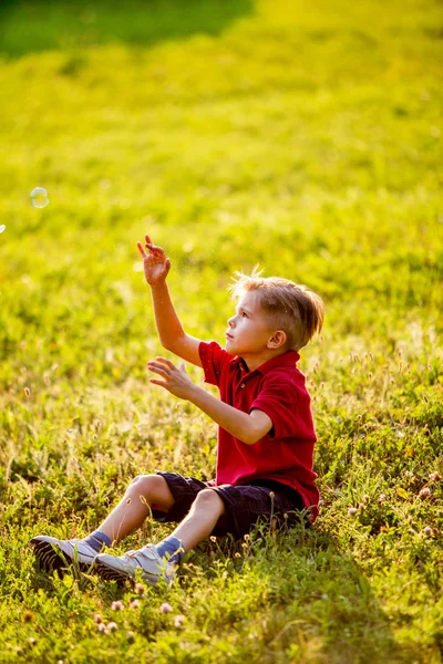 Мальчик, сидящий на зеленой траве, играет с мыльными пузырями — стоковое фото
