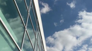 Mavi gökyüzü ve bulutlar güneşli bir günde cam endüstriyel bir binaya yansıyor.