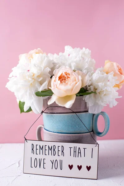 Sign with love words and tender summer roses and peonies flowers in vase against pink background. Selective focus. Still life. Postcard. Summer concept.