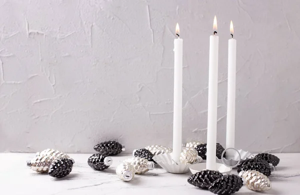 Scandinavian Christmas decorations. Candleholders with white burning candles, black, silver glass pinr cones on grey textured background.  Place for text. Postcard. Minimalistic style. Still life.