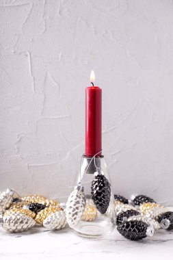 Noel süslemeleriyle dolu bir arka plan. Altın, siyah, gümüş renkli dekoratif koniler ve gri desenli arka planda yanan kırmızı bir mum taşıyan cam şamdan. İskandinav minimalist tarzı. Durgun hayat.