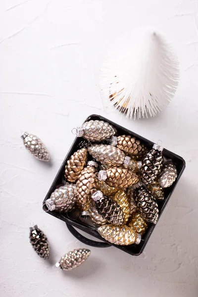 Christmas decorations. Metallic box with glass gold, black, silver decorative cones and white tree on white  textured background. Scandinavian minimalistic style. Still life. Place for text. Top view.