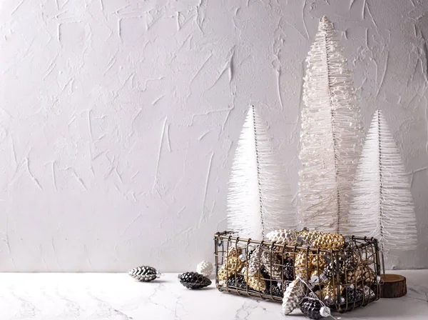 Christmas decorations. Metallic box with glass gold, black, silver decorative cones and white trees on white  textured background. Scandinavian minimalistic style. Still life. Place for text.