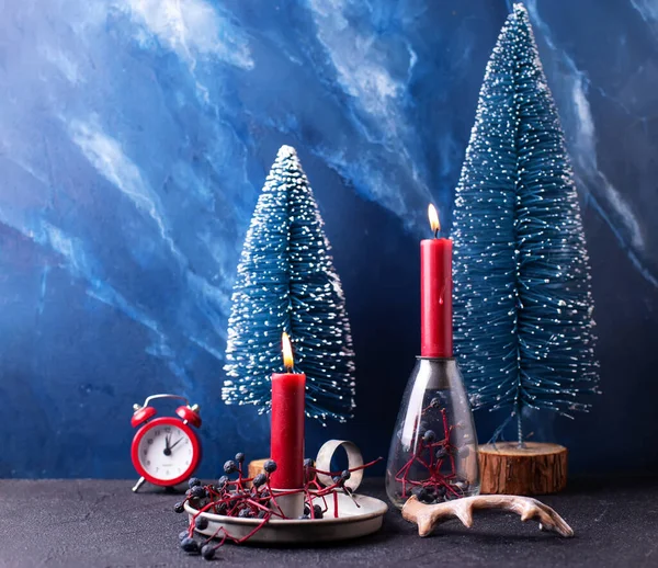 Bright Christmas  composition in red and blue colors. Red burning candles, wild blue berries, red clock and blue decorative trees against blue  textured wall. Still life. Place for text.