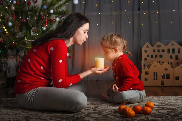 赤いセーターを着た若い幸せな母親と娘が家でろうそくを吹き消します。クリスマスムード、段ボールからDIYホーム装飾... — ストック写真