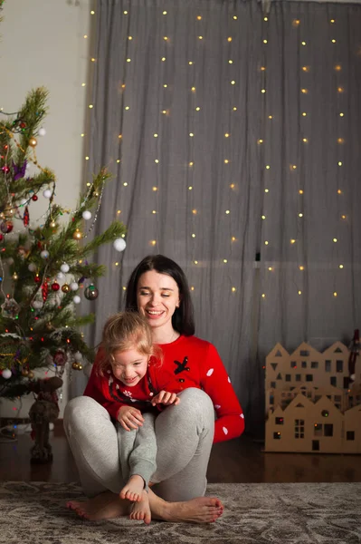 Kırmızı kazaklı genç mutlu anne ve kız evde oynaşıyor, sarılıyor, oynaşıyor. Noel havası, kartondan ev dekorasyonu.... — Stok fotoğraf