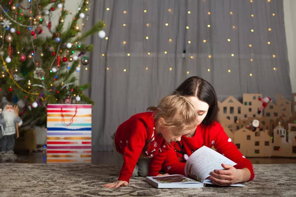 赤いセーターを着た若い幸せな母親と娘は自宅で本を読んでいます。クリスマスムード、段ボールからDIYホーム装飾... — ストック写真