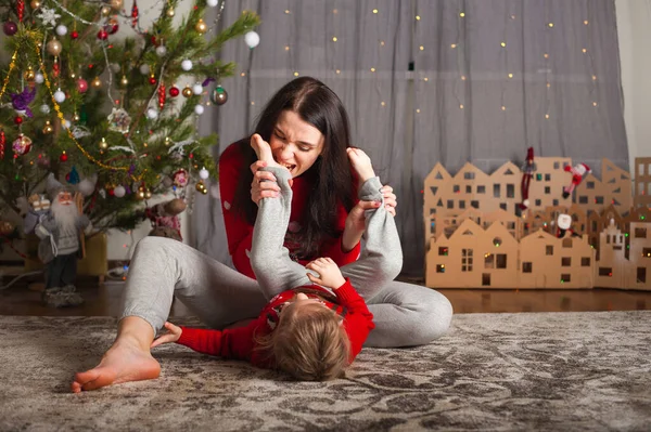 Kırmızı kazaklı genç mutlu anne ve kız evde oynaşıyor, sarılıyor, oynaşıyor. Noel havası, kartondan ev dekorasyonu.... — Stok fotoğraf