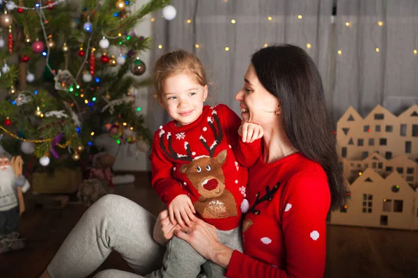 Kırmızı kazaklı genç mutlu anne ve kız evde oynaşıyor, sarılıyor, oynaşıyor. Noel havası, kartondan ev dekorasyonu.... — Stok fotoğraf