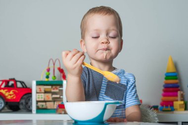 Küçük çocuk kaşıkla yer ve yiyecekle kaplıdır. Bebek maması, gıda alerjisi, kendini besleme.....