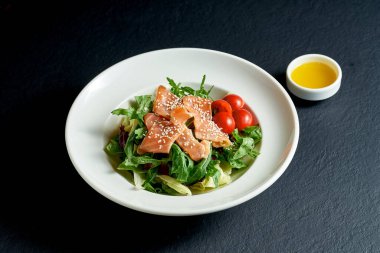 Siyah arka planda beyaz tabakta domatesli diyet salatası ve somon balığı.