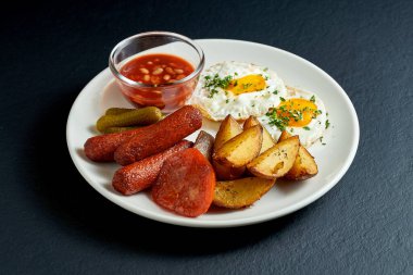 İngiliz kahvaltısı - patates, fasulye ve siyah arka planda beyaz bir tabakta çırpılmış yumurta, sosis.