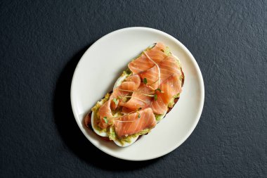 Siyah arka planda beyaz bir tabakta somon ve avokado kremalı tost.