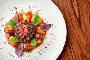 Fırında soğan ve sebzeli sığır filetosu, ahşap bir arka planda beyaz bir tabakta.