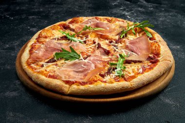 Pizza with jamon, arugula and sun-dried tomatoes on a black stone background. 45 degree angle view. Close-up, selective focus