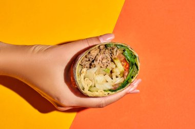 A hand holds a shawarma roll in a cut on a bright orange background
