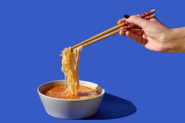 Yemek çubuğuyla kadın eli, erişte çorbasını mavi arka planda bir kaseye koyar.