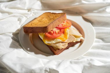 Kızarmış yumurta, tavuk ve domatesli tost sandviçi beyaz bir tabakta. Zor ışık.