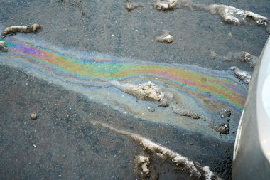 Asfalt bir yolda eriyen karın arka planına ve bir arabanın ön kısmına karşı bir yağ lekesi. Asfalt yolda renkli benzin lekeleri