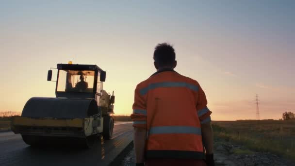 제복을 입은 한 남자 도로공사 가태 양 광선을 받고 있는 도로 로울러 로 걸어 간다. 뒤에서 쏘고 있어. 새로운 도로 건설. — 비디오