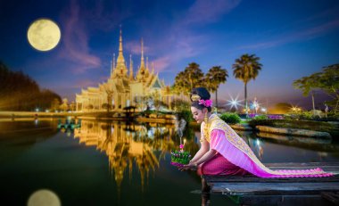 Beautiful Thai woman in Thai traditional costume on the wooden floor and holding floating basket (Krathong in Thai language) for floating in the red lotus lake in Loy Krathong Festival of Thailand. clipart