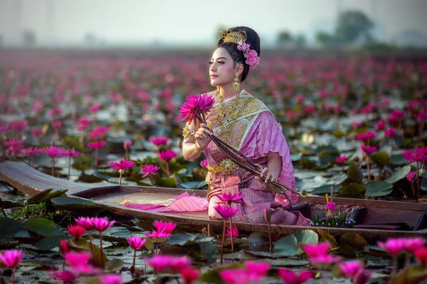 美丽的泰国女人穿着泰国传统服装坐在木船上 手里拿着红色的莲花在泰国洛城的红莲湖中 免版税图库照片