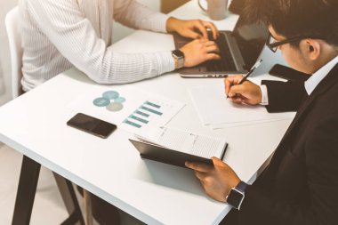 Masada oturan genç bir ofis çalışanı, bilgisayar kullanıyor. İki iş adamı konuşuyor.