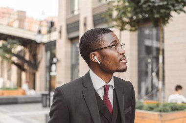 Siyah Afro-Amerikan iş adamının gözlüklü ve kulaklıklı portresi şehir binalarının arka planında duruyor.