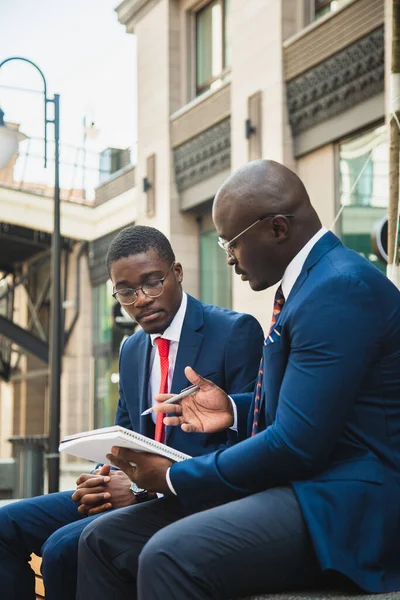 Rencontre amicale de deux hommes d'affaires afro-américains à la peau foncée à l'extérieur. Les partenaires s'assoient sur un banc et discutent d'un projet commun — Photo