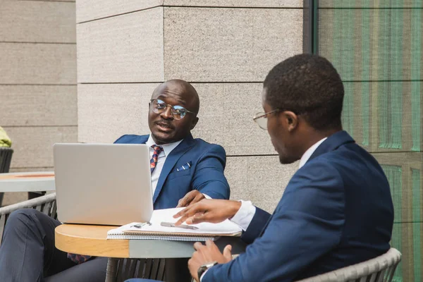 Deux hommes d'affaires afro-américains à la peau foncée en costumes et lunettes avec mallettes s'assoient à une table dans un café de la ville en plein air. — Photo