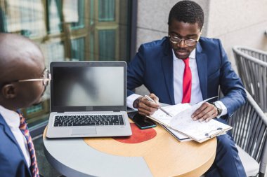 Siyah tenli, takım elbiseli ve çantalı iki Afro-Amerikan işadamı bir açık şehir kafesinde bir masada oturuyorlar..