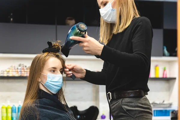 Il parrucchiere di donna di padrone in una maschera medica asciuga i capelli di ragazze con un asciugacapelli e pettini dopo essersi lavato in un salone di bellezza. Covid-19 pandemia e rimedi — Foto Stock