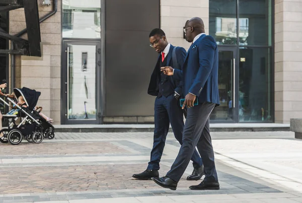 Dos hombres de negocios afroamericanos de piel oscura en trajes y gafas caminan al aire libre en la ciudad y hablan de negocios — Foto de Stock
