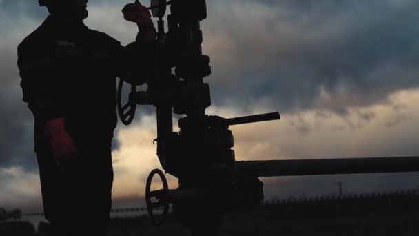 Работник в комбинезоне и шлеме проводит ремонт и обслуживание нефтяной скважины. Силуэт на фоне вечернего неба — стоковое видео