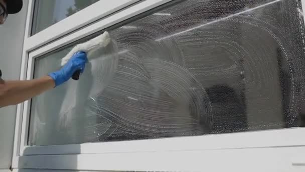 Un trabajador masculino profesional en overoles limpia hábilmente las ventanas y las enjabona con un equipo especial en cámara lenta. Servicios de limpieza — Vídeo de stock