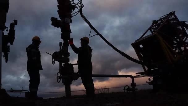 Los trabajadores en overoles y sombreros duros están llevando a cabo reparaciones y mantenimiento de un pozo de petróleo. Silueta en el fondo del cielo nocturno — Vídeos de Stock