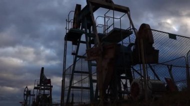 Enayi çubuğu pompalarının silueti çalışıyor ve akşam gökyüzünün zeminine yağ pompalıyor. Petrol kuyusu servisi