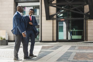 Koyu tenli iki Afro-Amerikalı takım elbiseli ve gözlüklü iş adamı şehirde yürüyüp iş konuşuyorlar.