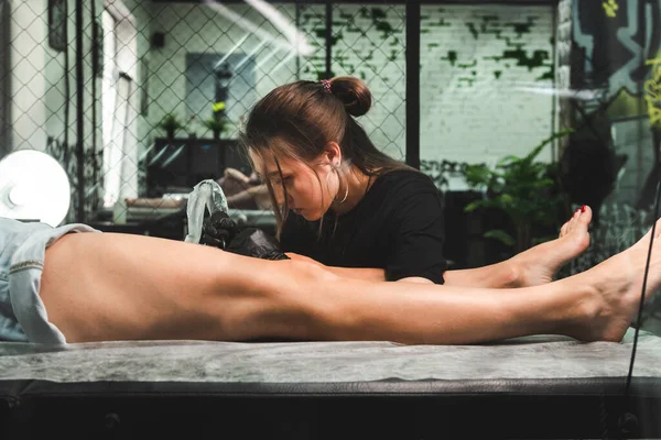 Un artista profesional del tatuaje hace un tatuaje en blanco y negro en una pierna de mujer, aplicando en tinta. El proceso de tatuar la piel. — Foto de Stock