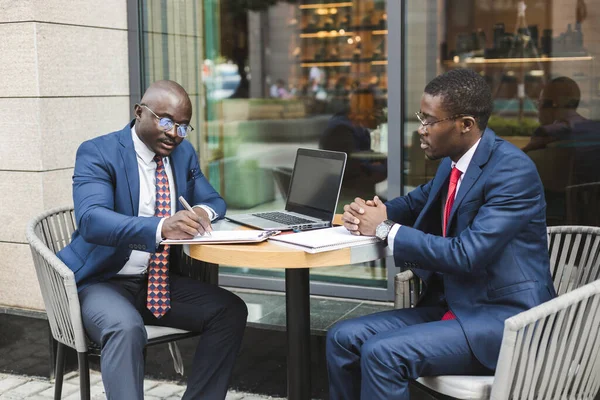 Deux hommes d'affaires afro-américains à la peau foncée en costumes et lunettes avec mallettes s'assoient à une table dans un café de la ville en plein air. — Photo