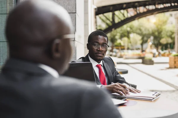 Två mörkhyade afroamerikanska affärsmän i kostym och glasögon med portföljer sitter vid ett bord på ett uteservering. — Stockfoto