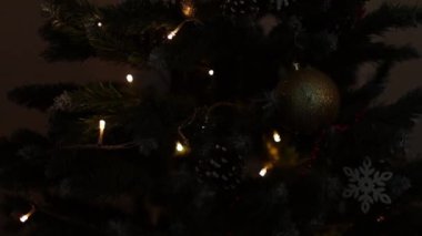 Noel ağacının dallarındaki Noel süslemeleri, arka plandaki çelenk ışıklarının sıcak ışıklarıyla titreşiyor..