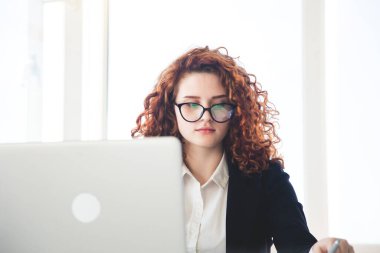 İş elbisesi ve gözlüklü güzel kıvırcık saçlı kadın ofiste dizüstü bilgisayarla çalışıyor.
